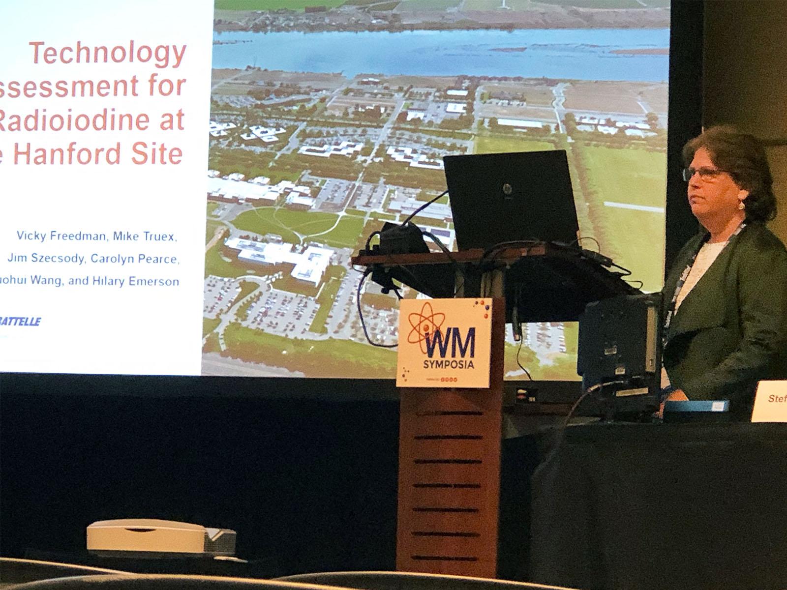 Vicky Freedman, a computational subsurface hydrologist, stands at a podium next to a big screen showing the title slide of her presentation
