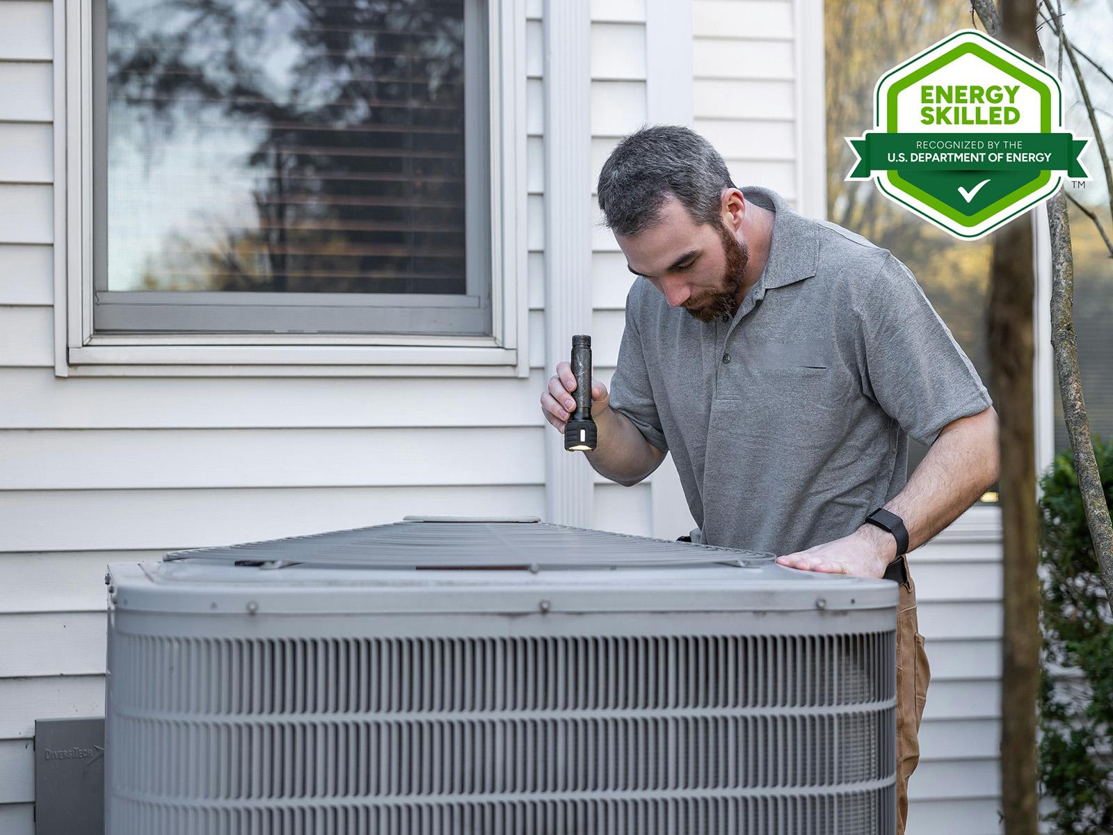 a man stands over an air conditioner, looking at it with a flashlight. In the corner there's a green and white DOE Energy Skilled badge