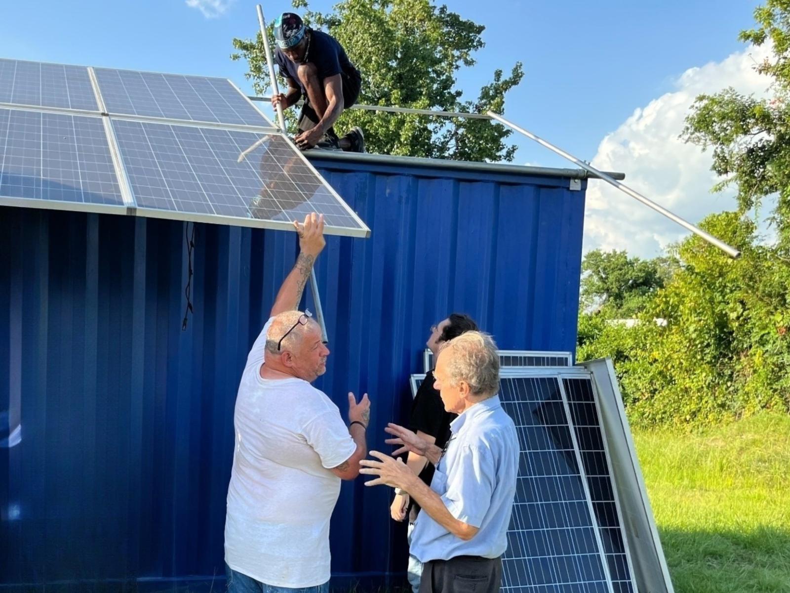 Two people install solar panels on the roof of a house while two others stand and converse near the house.