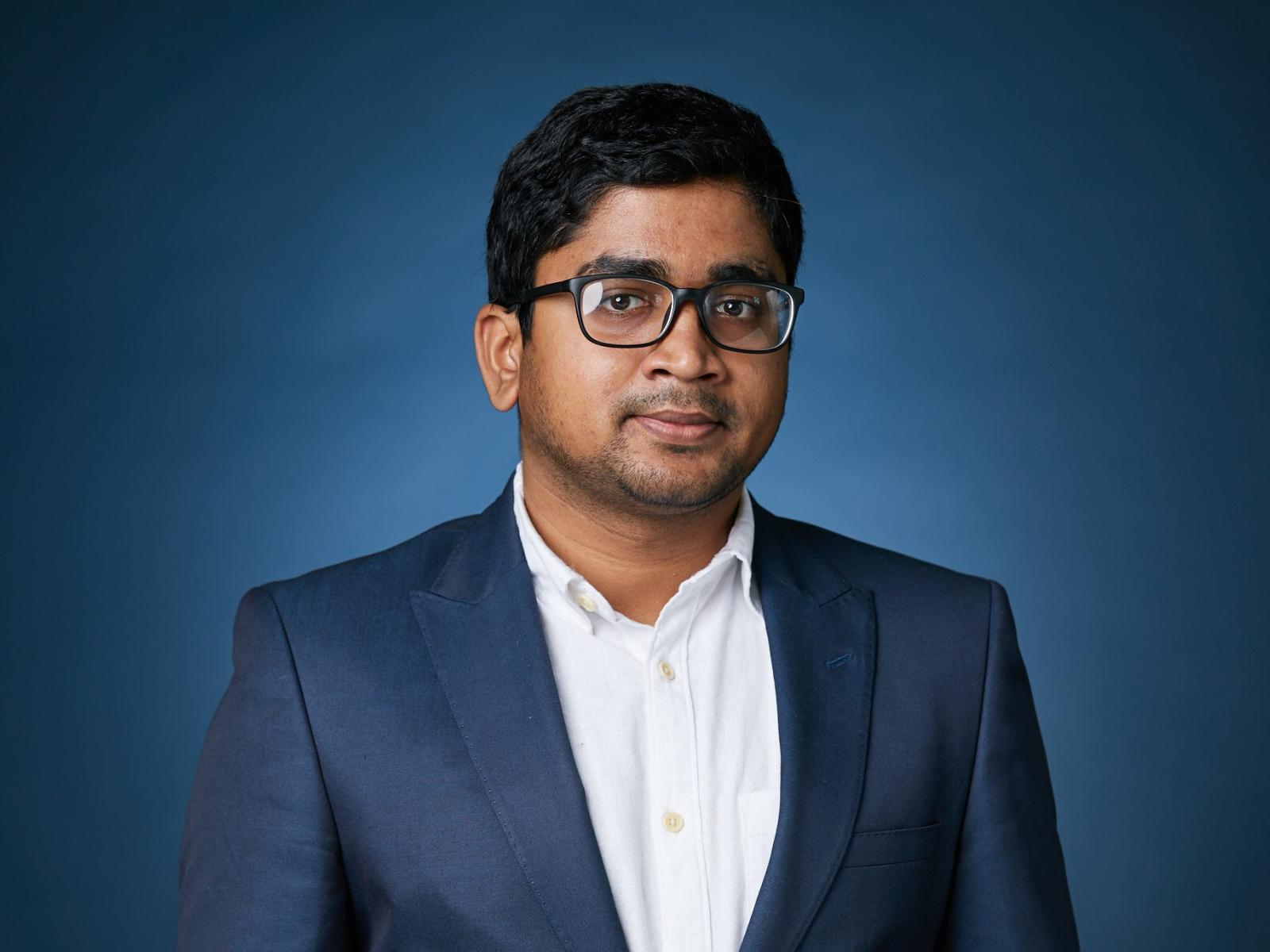 A person in a suit jacked and collared shirt with short hair and glasses pictured from the chest-up.