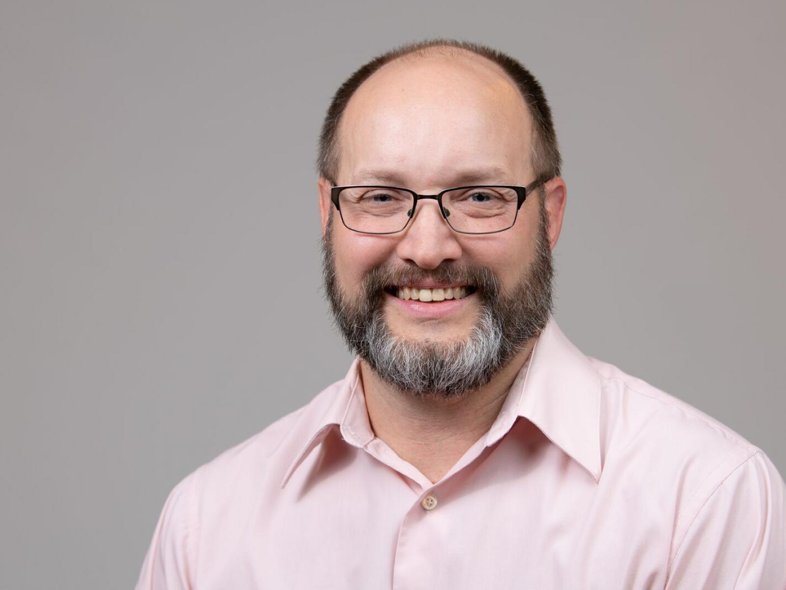 Photo of a man wearing a pink shirt and glasses smiling.