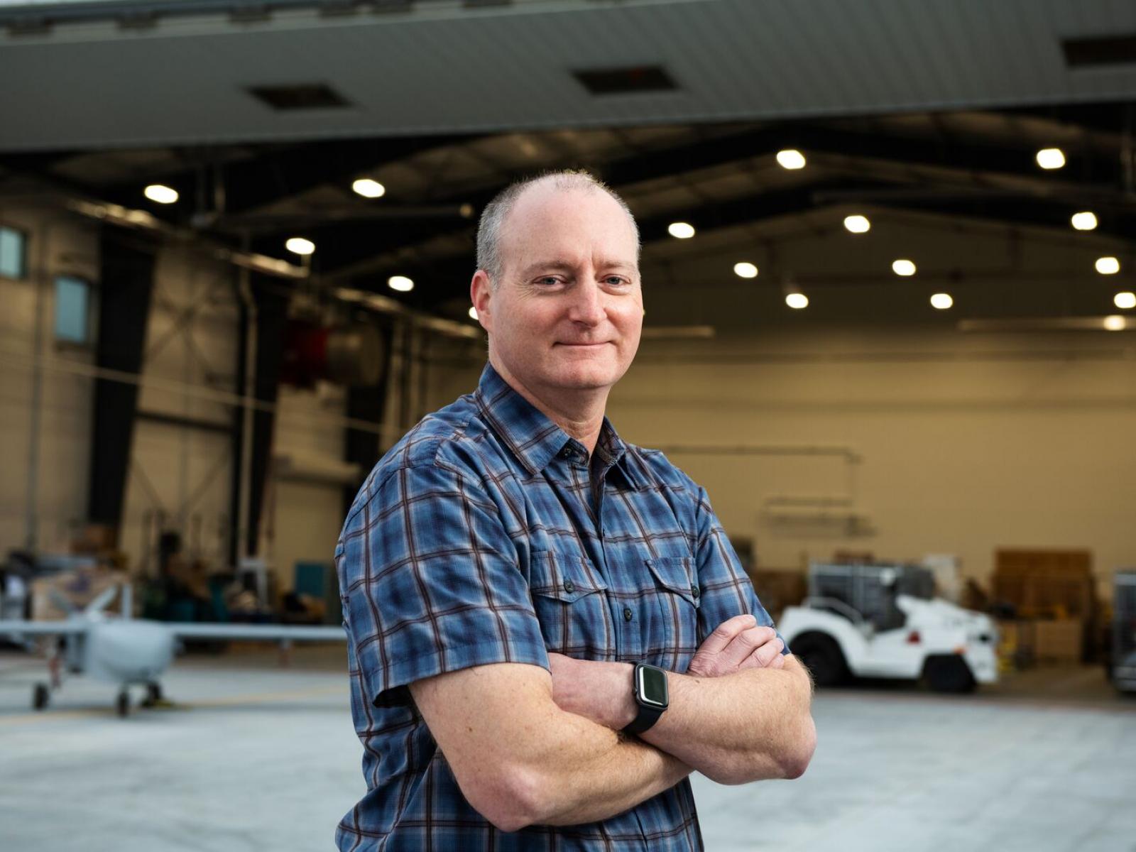 Photograph of smiling man in front of an aircraft hangar