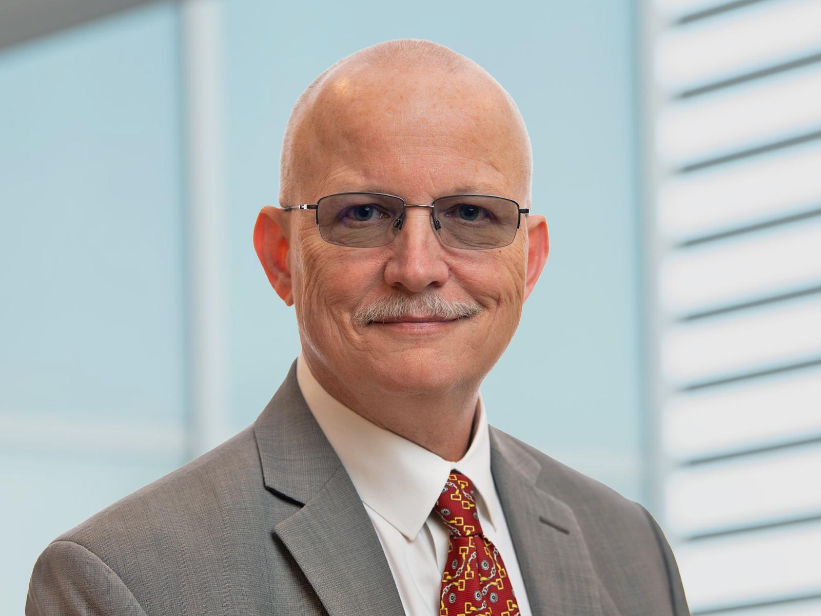 Mike Huyck pictured in front of a building, tight focus, facing the camera wearing glasses and a suit and tie