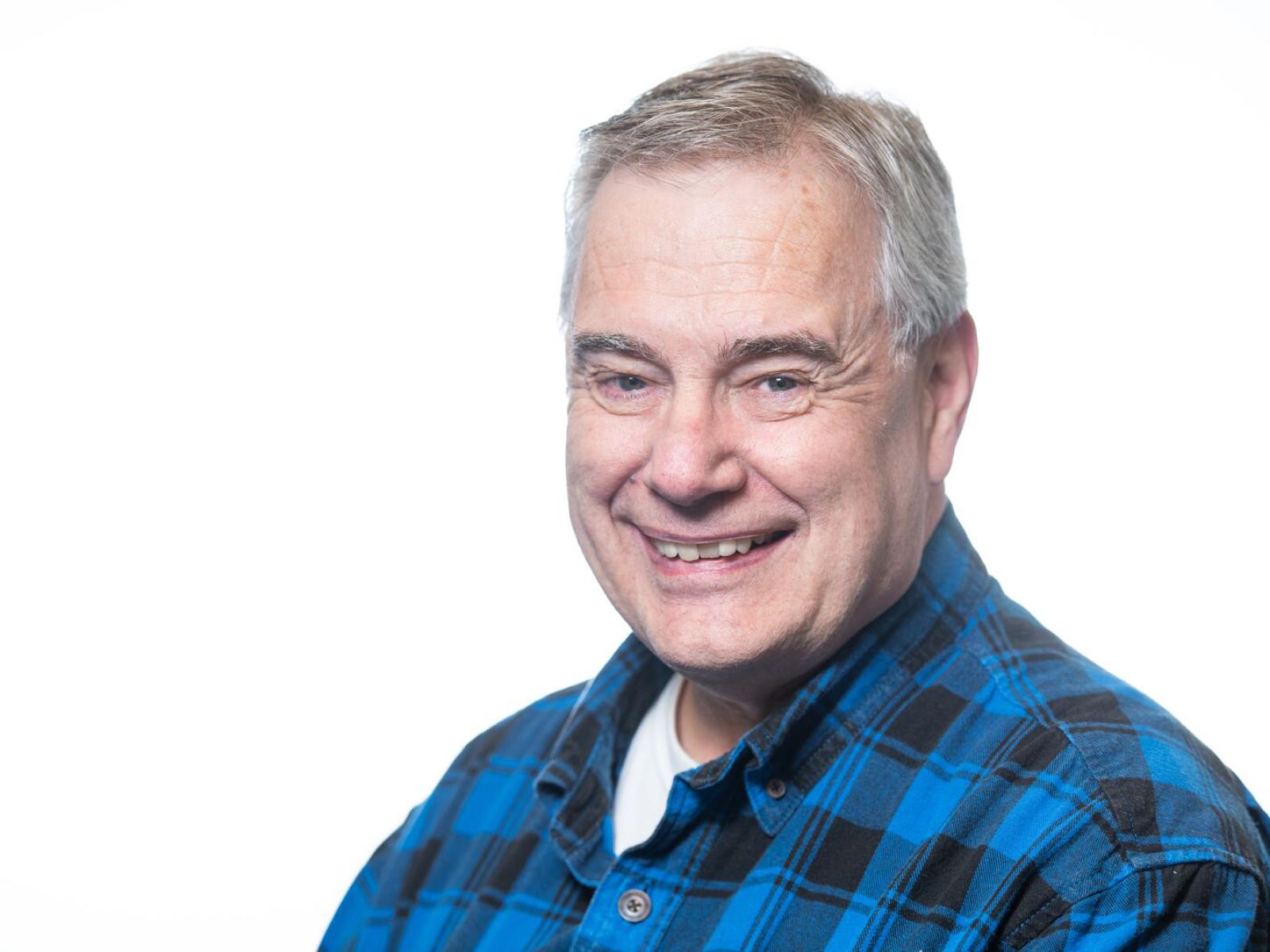 a smiling person with short gray hair pictured from the chest up, wearing a blue plaid collared shirt