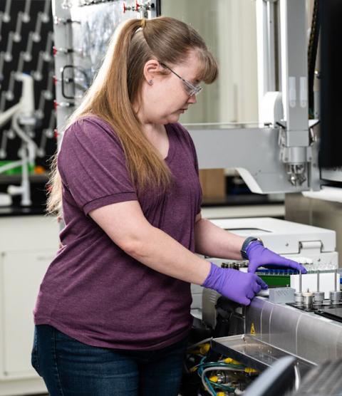 A woman stands next to a laboratory robot that has trays full of vials.