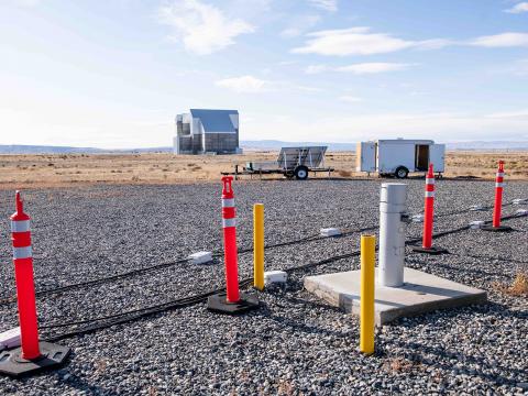 Black cables and orange cones are stretched across gravel in Hanford's 100 Areas, with the E4D white trailer and a Hanford cocooned building in the background