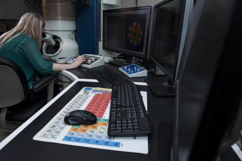 Female researcher views particles through eyepiece of large microscope while moving mouse with right hand to take scientific images.