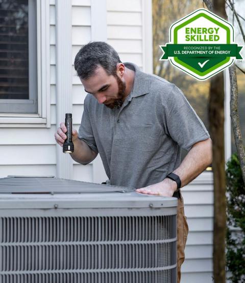 a man stands over an air conditioner, looking at it with a flashlight. In the corner there's a green and white DOE Energy Skilled badge