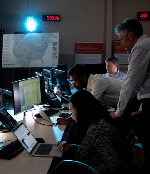 A woman sits at a desk, looking at a laptop. Two men stand behind her, also looking at the laptop. In the background are large screens showing graphs and grid simulations.
