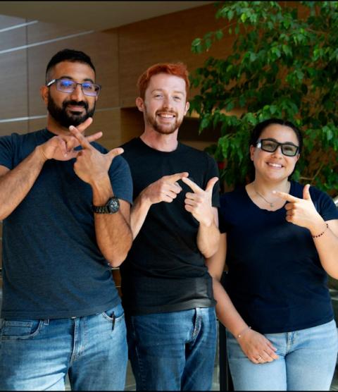 Three people stand in front of a PNNL entrance, each of them making part of the American Sign Language sign for "Pacific Northwest National Laboratory."