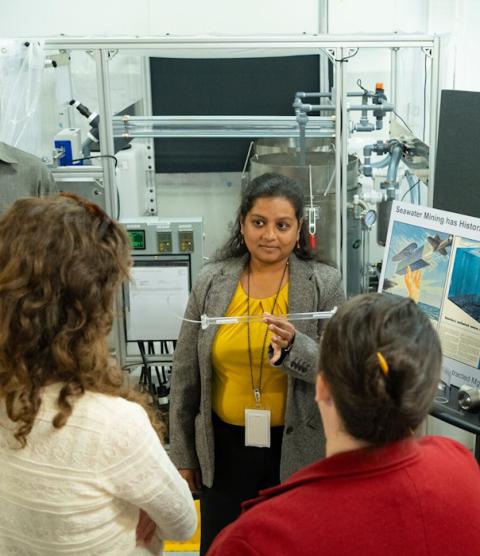 A woman in a grey blazer and yellow shirt holds up a plastic tube from a scientific instrument and shows it to a group of people.