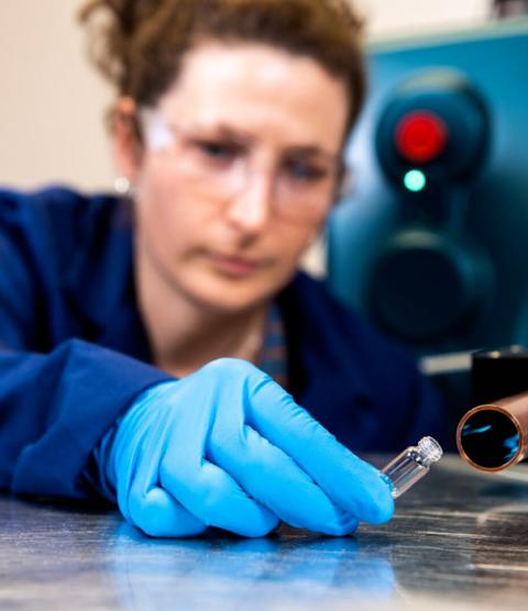 A researcher wearing safety clothing, gloves, and eyewear holding a small, open vial towards a sensor 