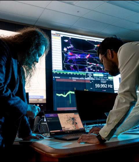 Two people stand over a desk looking at a laptop. Beyond them is a large screen covered in grid modeling data.