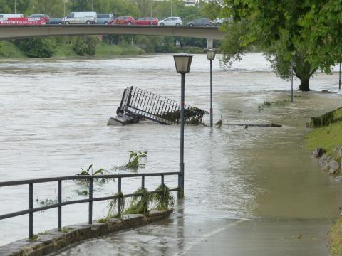 A rising river channel floods a paved walkway, submerging paths and nearby trees.