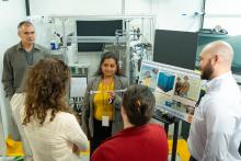 A woman in a grey blazer and yellow shirt holds up a plastic tube from a scientific instrument and shows it to a group of people.