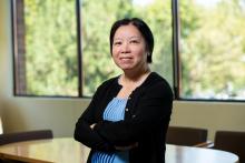 A woman with short black hair in black cardigan and periwinkle shirt smiling in front of a window in a conference room