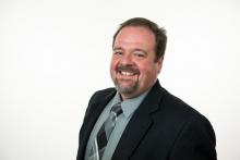 Smiling man with gray shirt, black sports jacket and multi-colored tie