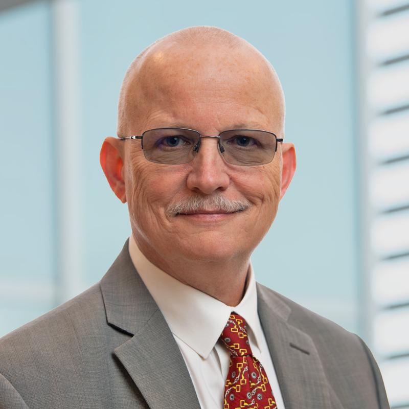 Mike Huyck pictured in front of a building, tight focus, facing the camera wearing glasses and a suit and tie