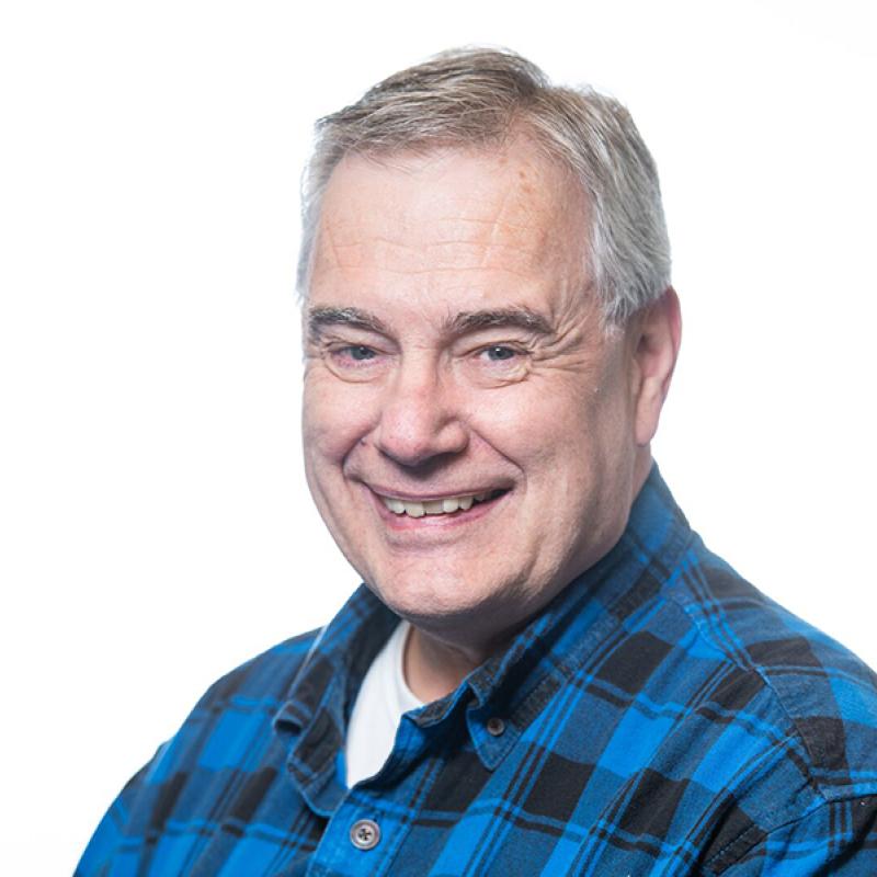 a smiling person with short gray hair pictured from the chest up, wearing a blue plaid collared shirt
