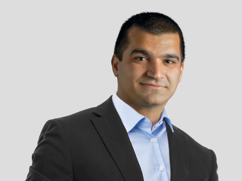Image of a slightly smiling middle eastern man with short hair wearing a dark suit and light blue shirt on a pale background.