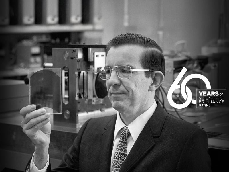 a man in a suit and tie and glasses holds up an early version of what would become a compact disc. The PNNL 60th anniversary logo is on the right.