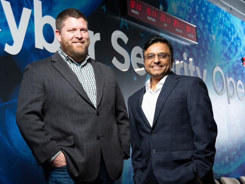 The photo shows Joseph Aguayo on the left and Mahantesh Halappanavar on the right, in front of a large blue wall that reads “Cyber Security Operations,” with a clock with black background and red digits at the top of the wall.