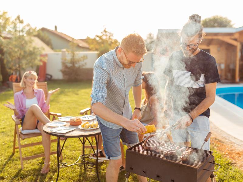 Four people in a backyard. Two sitting at a table and two manning a grill.