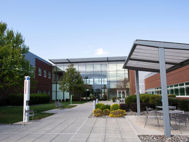 concrete tile walkway between brick buildings leading to a 2-story glass entryway flanked by bushes and trees