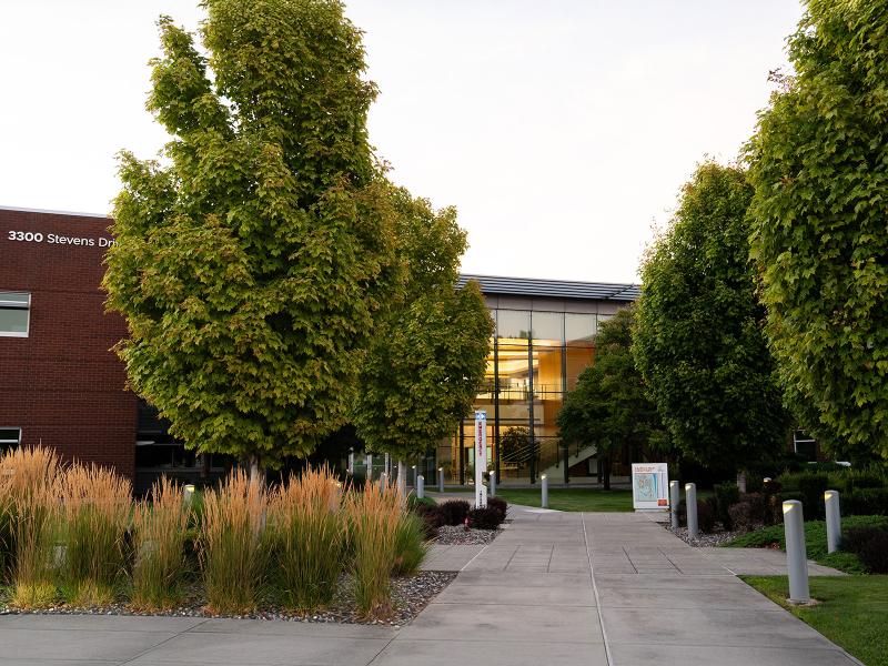 a tree- and brush-lined cement walkway leads to a glass-fronted bring building