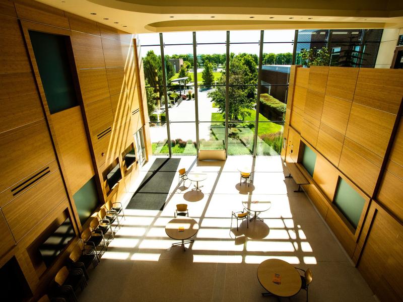 a sun-filled lobby with floor-to-ceiling two-story glass windows, round tables and chairs, and couches, looking out on a grassy area with trees and a concrete walkway