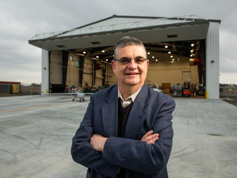 A man with short salt-and-pepper hair and glasses wearing a suit, standing in front of an open aircraft hangar with a small uncrewed aircraft behind him.
