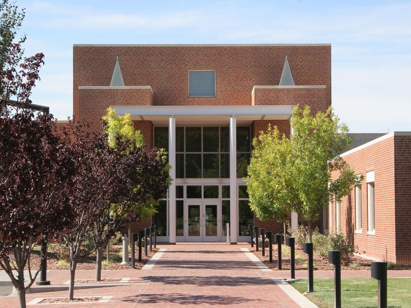 a tree-lined brick walkway leads to a tall, angular brick building with a two-story glass door entrance