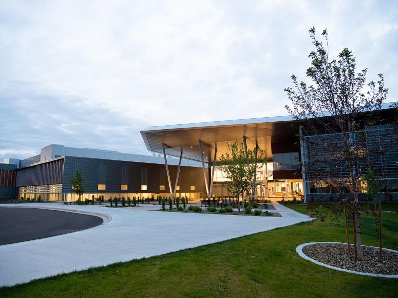a large brick and glass building in low light, illuminated from within,  with a concrete walkway and grass lawn in the foreground