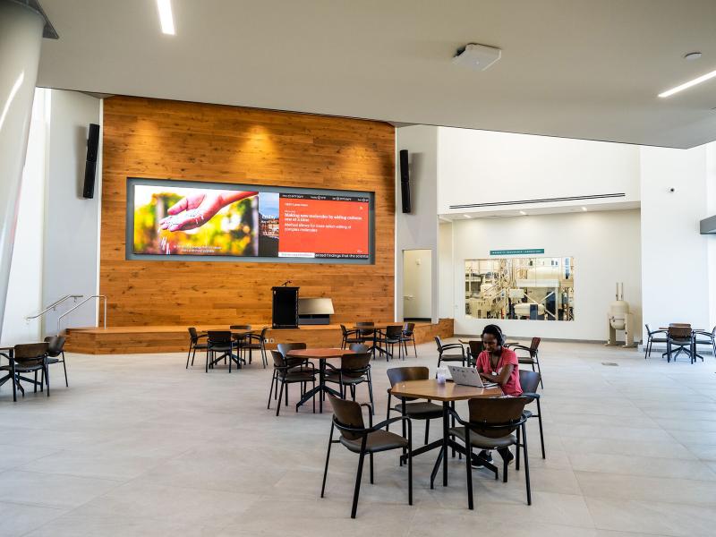 a bright interior lobby with round tables and chairs, a large flatscreen display on a rear wall, and windows looking into laboratory spaces