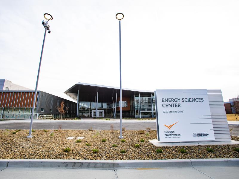 a rectangular white outdoor sign that reads "Energy Sciences Center" with a large brick and glass building in the background