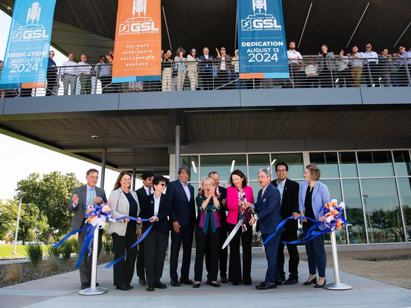 A group of people in professional dress stand together, smiling and laughing, after cutting a ribbon to dedicate a new building