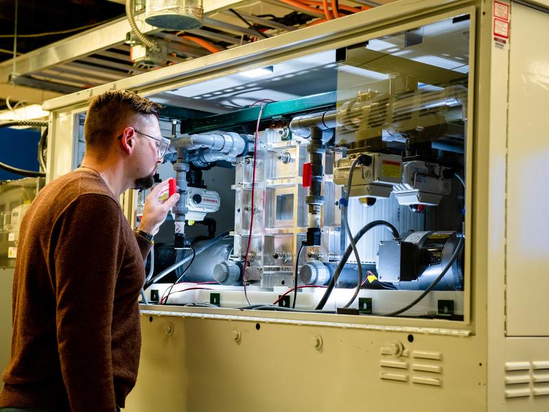 A researcher in protective eyewear observes a piece of equipment through a glass window