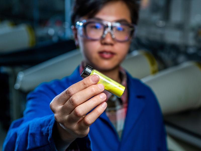 A researcher in a blue lab coat and protective eyewear holds a vial of a bright yellow powder toward the camera