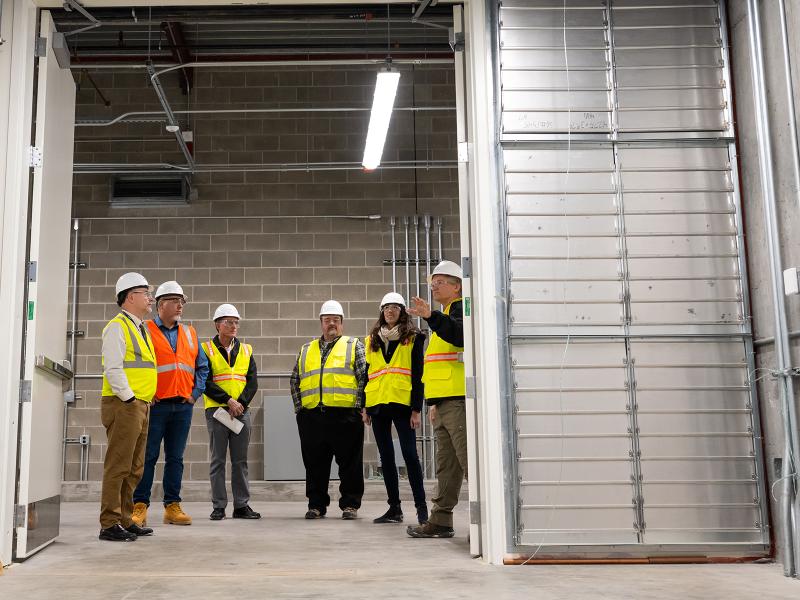 A group of professionally-dressed people in yellow reflective vests and hard hats examines a warehouse facility
