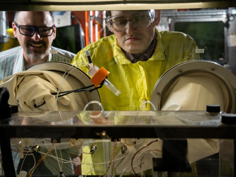 A scientist holds a vial using a set of tongs and rubber gloves. Another scientist watches him from behind.