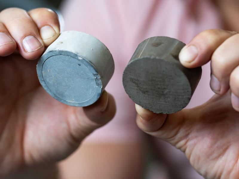 A researcher holds up two samples: on the left, a cylinder of cement made with traditional supplementary materials; on the right, a cylinder of cement made with algal biochar.