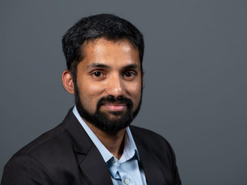 Asian man with short hair a trimmed beard slightly smiles at the camera while wearing a dark suit and blue shirt with its collar unbuttoned, all on a dark background