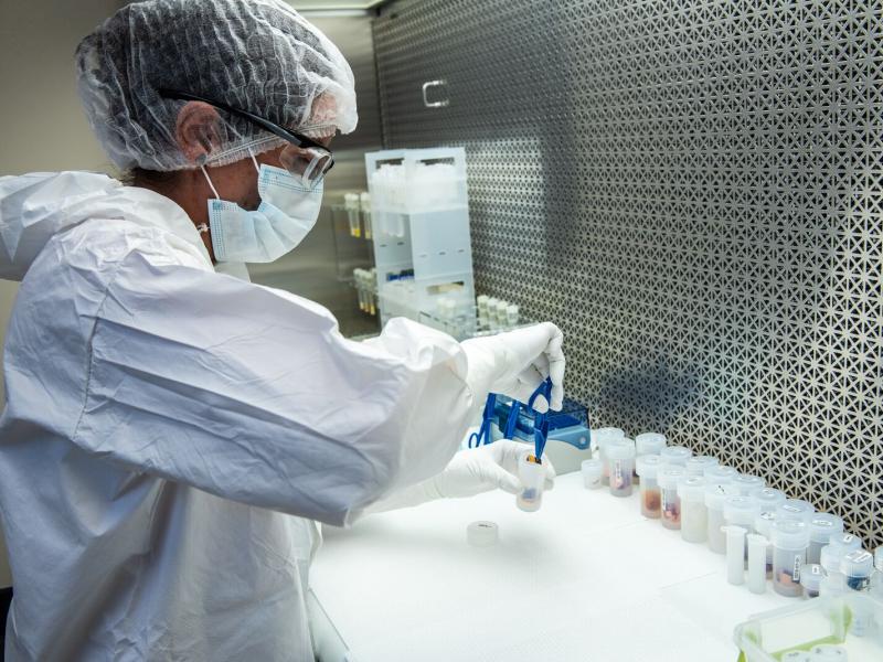 Woman in full PPE/clean room garb, grabbing samples at a bench
