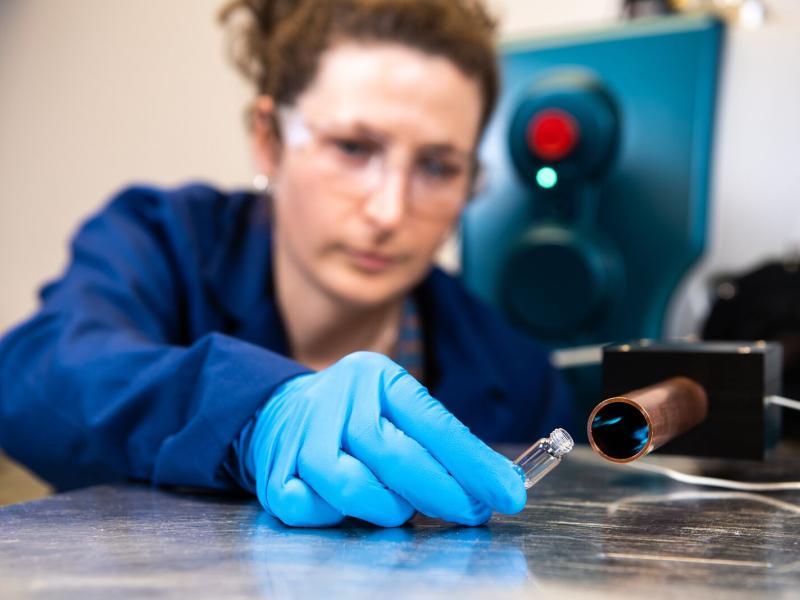 A scientist wearing protective clothing and eyewear holds a small glass tube at the entrance of a metal tube, collecting vapor.
