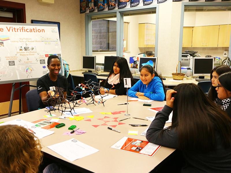STEM Ambassador Charmayne Lonergan at a table with students depicting a traditional on-site STEM outreach experience