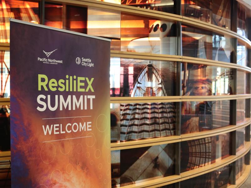 Photograph shows a sign that says "ResiliEX Summit; Welcome" in front of windows looking into a conference room.