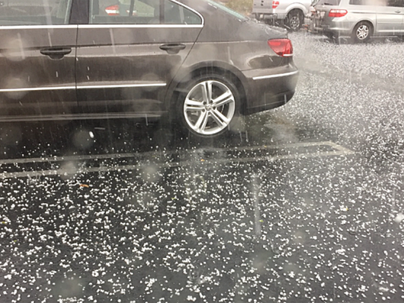 Photograph of a parking lot with cars during a hailstorm. There is a black sedan featured prominently with lots of hail on the black asphalt. The image is streaky from moving hail falling in front of the camera.
