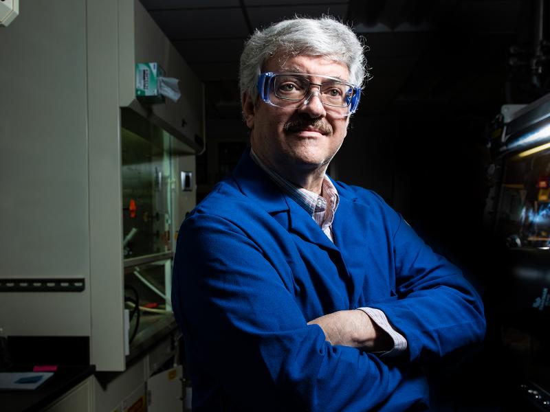 White man with gray hair, a moustache, and glasses wearing a cobalt blue lab coat standing with his arms crossed in a dark laboratory.