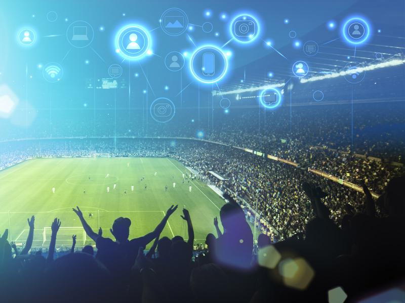 Photo of attendees watching two soccer teams compete in a sports stadium. Overlaying this photo is a digital web with icons representing mobile devices, user accounts, camera, laptops, text messages, and internet connectivity all connected together.