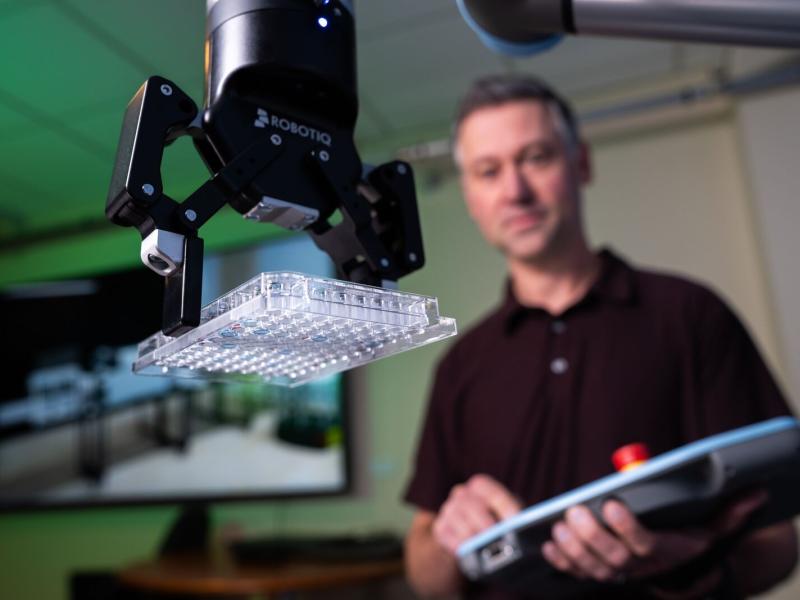 A researcher with a control device standing behind a robotic arm holding a lab sample tray. 